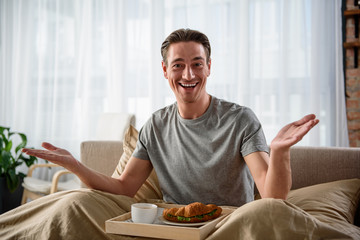 Waist up portrait of cheerful guy looking at camera with smile. He is sitting on bed with food on tray. Concept of joy