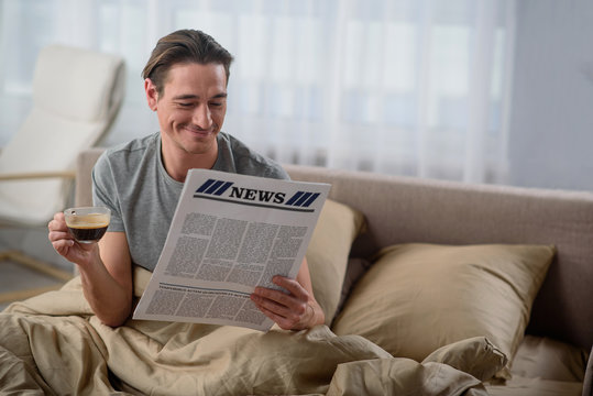 Waist up portrait of happy guy reading the newspaper. He is holding a cup of coffee and sitting in bedroom. Copy space in right side
