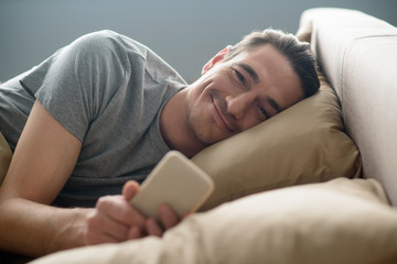 Happy guy looking at camera with smile. He is lying on bed and holding mobile phone