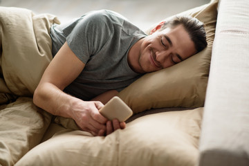 Satisfied man is lying in bed and looking at the mobile phone with smile in the morning
