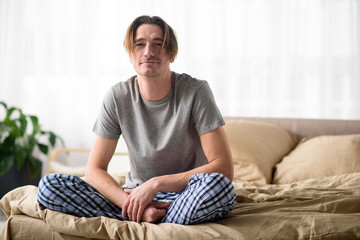 Good morning. Full length portrait of happy man sitting on bed and looking at camera with calmness. Relaxation concept