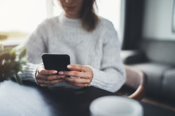 Closeup image of young smiling female blogger using modern smartphone device while sitting at cozy home interior, communication and social networks concept, flare light effect