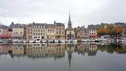 Fototapeta premium HONFLEUR, FRANCE - Scenic view of church and saling boats in old medieval harbor, beautiful buildings in historic city center, people are relaxing at terraces along the harbour in old town