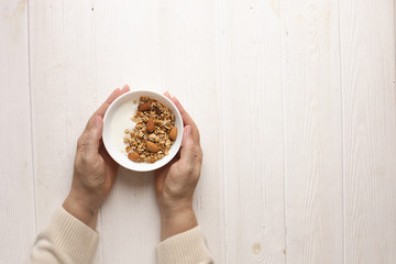 Woman eating homemade granola breakfast cereals with mixed nuts, oats and Greek yogurt in bowl. Female hands holding healthy vegetarian food with cashew, almond, hazelnut. Morning light, copy space.