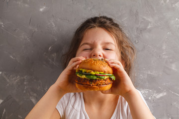 The little girl is eating a healthy baked sweet potato burger with a whole grains bun, guacamole, vegan mayonnaise and vegetables. Child vegan concept, gray background. - obrazy, fototapety, plakaty