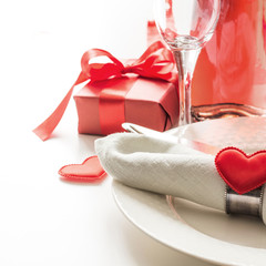Valentines day dinner with table place setting with red gift, a bottle of champagne, heart ornaments with silverware on white. Close up. Valentine's card.