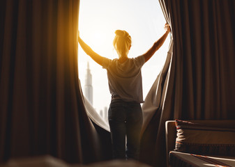 happy woman stretches and  opens curtains at window in morning