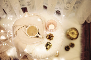Hot chocolate, a cup of cappuccino on a fur chair. Warm scarf, cones, lights around. Cozy winter evenings