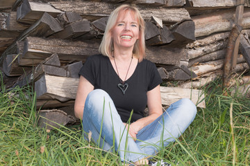 Blonde Frau sitzt lachend vor Brennholzstapel im Gras