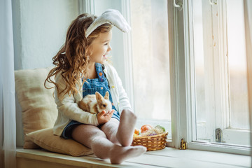 Cute happy little child girl is wearing bunny ears on Easter day sitting on window sill holding her friend little colorful rabbit