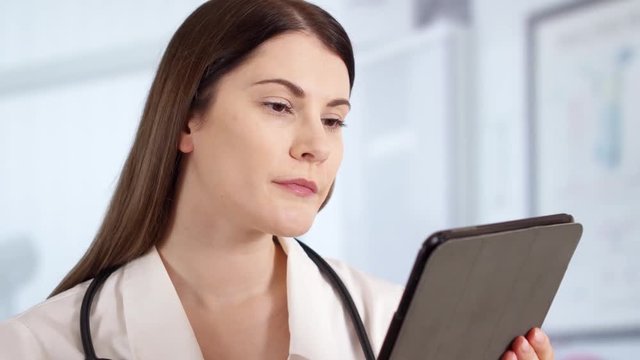 Portrait of smiling confident professional female doctor with stethoscope standing in hospital room using tablet. Woman physician at work. Health care concept