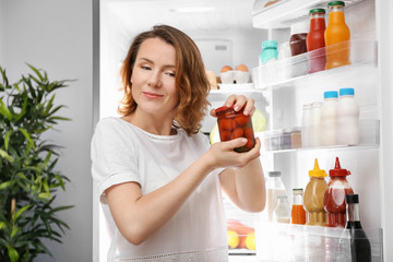 Beautiful woman opening jar with tomatoes near refrigerator