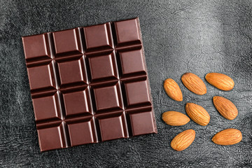 Dark, bitter chocolate bar and almond nuts on black leather texture background, top view