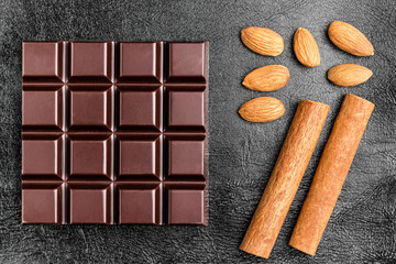 Dark, bitter chocolate bar, almond nuts and two cinnamon sticks on black leather texture background, top view