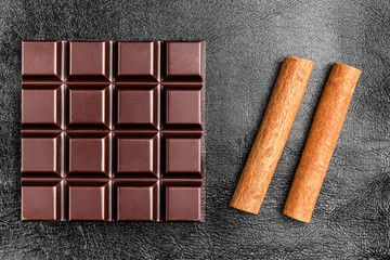 Dark, bitter chocolate bar and two cinnamon sticks on black leather texture background, top view