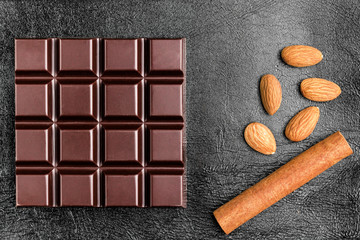 Dark, bitter chocolate bar, almond nuts and cinnamon stick on black leather texture background, top view