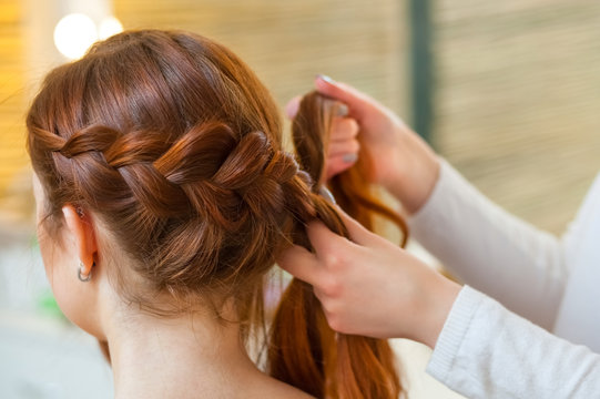 Beautiful girl with red hair, hairdresser weaves a  braid close-up, in a beauty salon. Professional hair care and creating hairstyles.