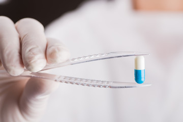 Hand in latex glove holding tweezers with blue and white capsule. Pharmaceutical research.
