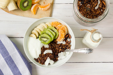 A bowl of chocolate cereal with yogurt and seasonal fruits in a white bowl, top view, ingredients in the background