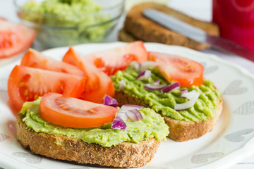 Fototapeta na wymiar Whole wheat bread with green avocado cream and fresh tomatoes on white plate. Ingredients in the background