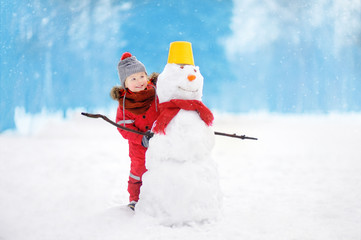 Little boy in red winter clothes having fun with snowman in snowy park