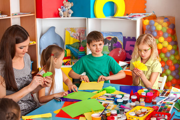 School children with scissors in kids hands cutting paper with teacher in class room. Group of children in an orphanage. Preparation for origami exhibition. Large group girls and boys together.