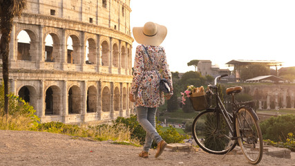 Young woman tourist with bycicle takes pictures of the colosseum in rome at sunset with smartphone....
