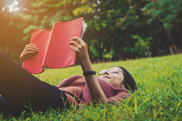 Young woman wearing glasses enjoy reading a red book lying on green grass