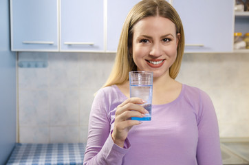 Beautiful woman, holding glass of fresh water in kitchen, looking at camera and smiling 