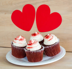 4 chocolate cupcakes with white frosting and red sprinkles on a wooden table with 2 red hearts above them