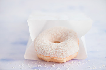 Close-up of a white donut with coconut topping on a light background inside paper delivery packaging. High key food photography.