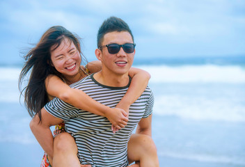  beautiful Asian Chinese couple with boyfriend carrying woman on her back and shoulders at the beach smiling happy in love enjoying holidays