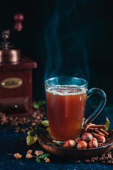 Steaming latte glass with coffee grinder, spices, nuts and coffee beans on a dark background. Action drink photography. Brewing coffee concept.