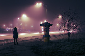 A lonely woman walking along the night street, lit by street lights and car headlights.