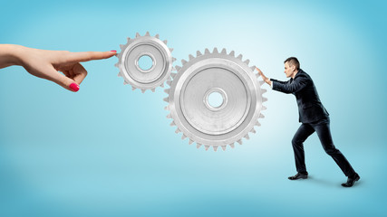 A small businessman pushes at a large gear interlocked with a small one while a female finger stops their movement.