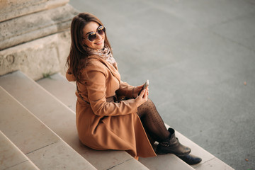 Stylish girl in a brown coat and a scarf with glasses. Using a phone in paris france. Makeup