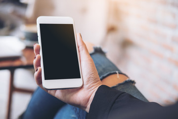 Mockup image of a woman's hand holding white mobile phone with blank black desktop screen on thigh in cafe