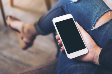 Mockup image of a woman's hand holding white mobile phone with blank black desktop screen on thigh in cafe