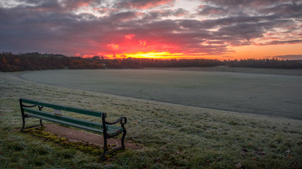 Fototapeta na wymiar A beautiful early morning sunrise over an open frosty field parkland with an empty bench seat in the foreground