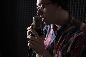 Close-up portrait of a young man singing with closed eyes in glasses and a hat holding a microphone in his hand.