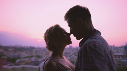 Cute lovers gently hugging, standing on roof, beautiful sunset city background
