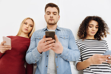 So serious. Attractive blonde girl holding her gadget and leaning on wall while looking at telephone of her friend