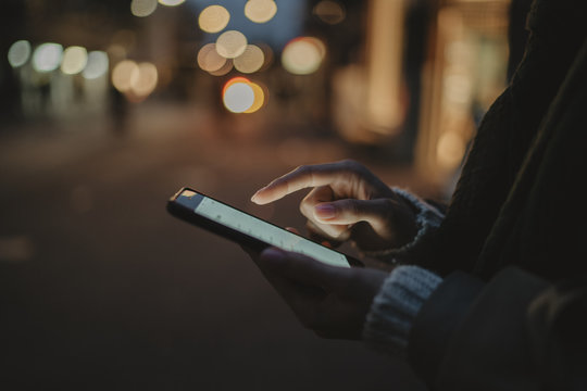 Cropped image of female hands using modern smartphone at night, bokeh light on the background