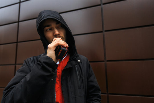 Vape man. Portrait of a bearded handsome young white guy in the hood vaping an electronic cigarette opposite the futuristic urban background.