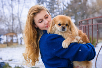 Woman pet owner with blue coat and golden hair walking during winter with her dog. Young lady  with pekingese dog walking on the snowy field. Tender feeling with a dog 