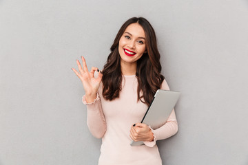 Attractive female with beautiful smile holding silver notebook gesturing OK sign over grey wall