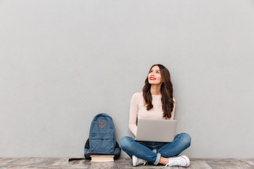 Horizontal image of female looking upward while reading or communicating online using silver laptop sitting with legs crossed on the floor over gray wall - obrazy, fototapety, plakaty
