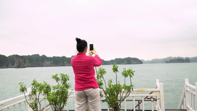 The woman in a pink jacket takes the picture. The woman travels by the tourist boat around a fine bay of Vietnam. The smiling woman takes the picture on the deck of the tourist boat. Vietnam.