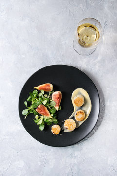 Fried scallops with lemon, figs, sauce and green salad served on ceramic black plate with glass of white wine over gray texture background. Top view, space. Plating, fine dining