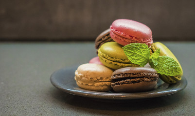 Assortment of color macaroon cookies on plate, decorated with mint leafs, dark background. green pistachios, brown chocolate, pink strawberry and creme vanilla almond cakes. copyspace for text